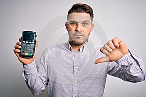 Young business man with blue eyes holding dataphone payment terminal over isolated background with angry face, negative sign