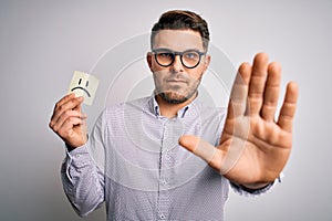 Young business man with blue eyes holding paper note with sad face as unhappy message with open hand doing stop sign with serious