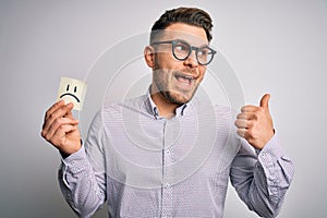 Young business man with blue eyes holding paper note with sad face as unhappy message pointing and showing with thumb up to the