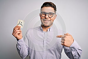 Young business man with blue eyes holding paper note with sad face as unhappy message with surprise face pointing finger to