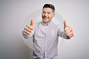 Young business man with blue eyes standing over isolated background approving doing positive gesture with hand, thumbs up smiling