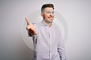 Young business man with blue eyes standing over isolated background smiling friendly offering handshake as greeting and welcoming