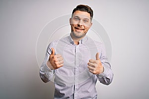 Young business man with blue eyes standing over isolated background success sign doing positive gesture with hand, thumbs up