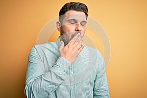 Young business man with blue eyes wearing elegant green shirt over yellow background bored yawning tired covering mouth with hand