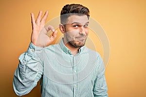 Young business man with blue eyes wearing elegant green shirt over yellow background smiling positive doing ok sign with hand and