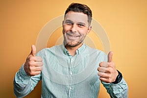 Young business man with blue eyes wearing elegant green shirt over yellow background success sign doing positive gesture with