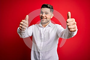 Young business man with blue eyes wearing elegant shirt standing over red isolated background approving doing positive gesture