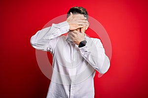 Young business man with blue eyes wearing elegant shirt standing over red isolated background Covering eyes and mouth with hands,