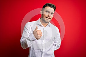 Young business man with blue eyes wearing elegant shirt standing over red isolated background doing happy thumbs up gesture with