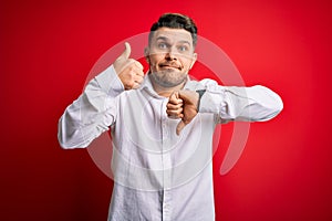 Young business man with blue eyes wearing elegant shirt standing over red isolated background Doing thumbs up and down,