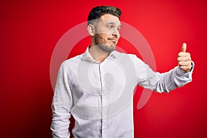 Young business man with blue eyes wearing elegant shirt standing over red isolated background Looking proud, smiling doing thumbs