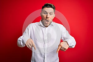 Young business man with blue eyes wearing elegant shirt standing over red isolated background Pointing down with fingers showing