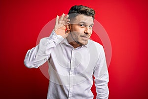 Young business man with blue eyes wearing elegant shirt standing over red isolated background smiling with hand over ear listening
