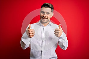 Young business man with blue eyes wearing elegant shirt standing over red isolated background success sign doing positive gesture