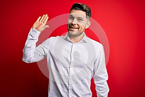 Young business man with blue eyes wearing elegant shirt standing over red isolated background Waiving saying hello happy and