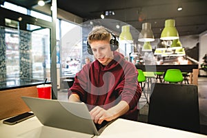 Young business man in casual clothing and headphones, sitting at a cafe, using a laptop, looking at the screen and smiling