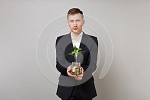 Young business man in classic black suit shirt holding pile gold coins in glass jar with green plant sprout isolated on