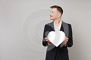 Young business man in classic black suit, shirt looking aside holding white heart with copy space isolated on grey
