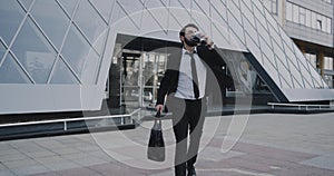 Young business man finished his work day comes out from modern building holding coffee and briefcase