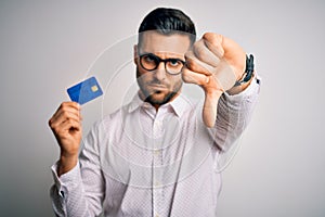 Young business man holding credit card over isolated background with angry face, negative sign showing dislike with thumbs down,