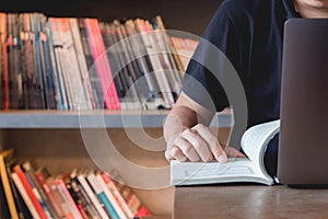 Young business man learning stock trading. Man sitting in library reading book, studying book  using laptop making research.