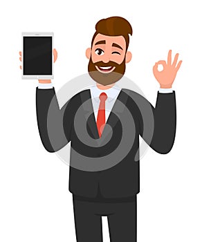 Young business man showing/holding blank screen of tablet computer display and gesturing/making okay, ok sign, while winking eye.