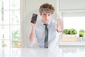 Young business man showing smartphone screen at the office with open hand doing stop sign with serious and confident expression,