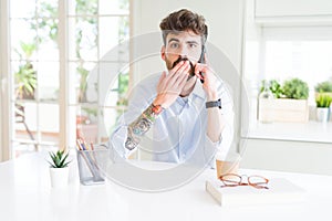 Young business man talking on smartphone cover mouth with hand shocked with shame for mistake, expression of fear, scared in