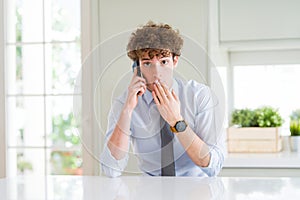 Young business man talking on smartphone at the office cover mouth with hand shocked with shame for mistake, expression of fear,