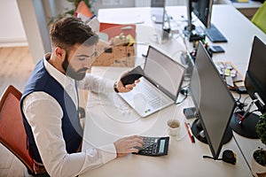 Young business man using calculator at his desk in office, processing data, holding cell phone in front of computer. business ,