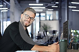 Young business man is using laptop while sitting in the office, looking at camera, smiling