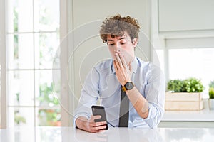 Young business man using smartphone at the office cover mouth with hand shocked with shame for mistake, expression of fear, scared