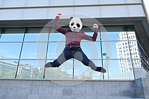 Young business man wearing a panda head mask, jumping in front of the a corporate building
