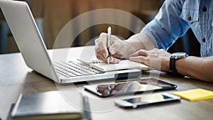 Young business man working in bright office, using laptop, writing notes.