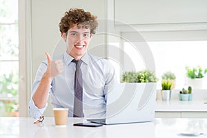 Young business man working with computer laptop at the office happy with big smile doing ok sign, thumb up with fingers, excellent