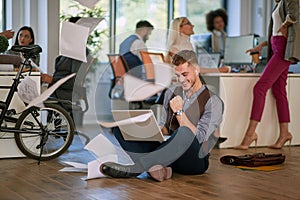 Young business man with fist up, looking at his laptop, sitting on a floor ina a modern, casual, open space office. casual