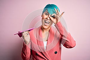 Young business woman with blue fashion hair holding paper plane as travel symbol with happy face smiling doing ok sign with hand