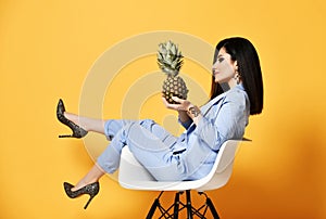 Young business woman in blue official suit sits in armchair sideways and looks with interest at pineapple she holds