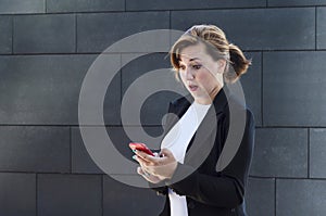 Young business woman in a business suit looks in surprise at the phone. girl, lady receives and reads good news.