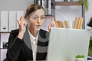 Young business woman crossed fingers looking at laptop screen asking for good luck news win victory