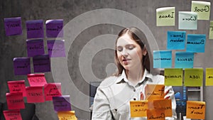 Young business woman is doing brainstorm using stickers on the glass. Office worker is thinking about strategy, business