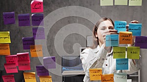 Young business woman is doing brainstorm using stickers on the glass. Office worker is thinking about strategy, business