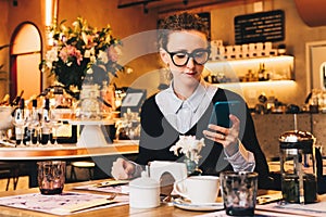 Young business woman in glasses sits in cafe at table, uses smartphone. On table is cup of coffee. Girl working