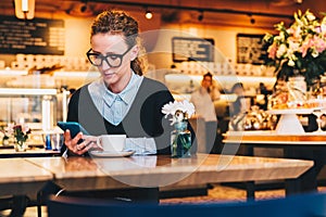 Young business woman in glasses sits in cafe at table, uses smartphone. On table is cup of coffee. Girl working