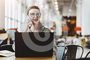 Young business woman in glasses sitting at table in cafe, talking on cell phone while working on laptop.Student learning