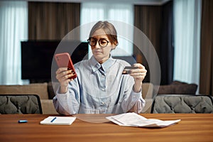 Young business woman hold bank credit card look at phone screen paying bills taxes online by easy banking payment system