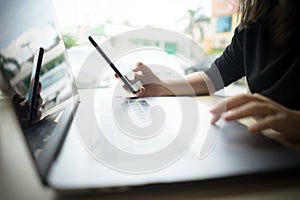 Young business woman holding mobile phone and typing on laptop computer keyboard in coffee shop