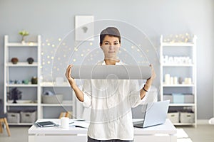 Young businesswoman holding yoga mat in hands standing in cozy office and looking at camera