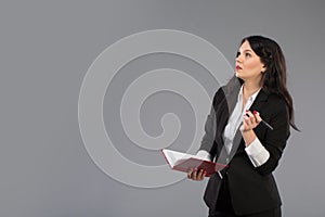 Young business woman with notepad and pen close to her chin. Pensive business lady writing on clipboard standing over