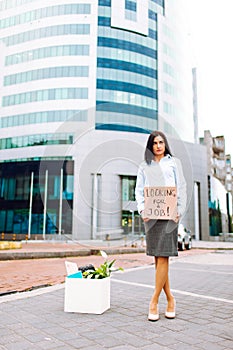 Young business woman in office staying near buisness centre with a box of her office supplies and cardbox looking for a job.